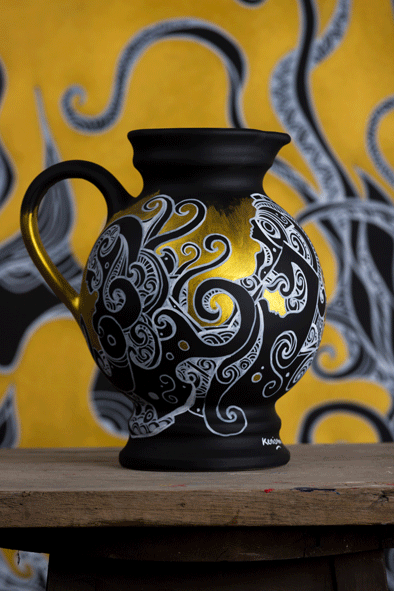 A big black jug that contains a beautiful elegant hand drawn artwork with acrylic paint by artist kennetn kenisman. The gold paint gives the artwork a feeling of depth. The artwork on the object depicts a nature scene, with curls, waves, sea or nature elements and intricate details. Also silhouette figures wearing an ancient helmet. The composition is well balanced.