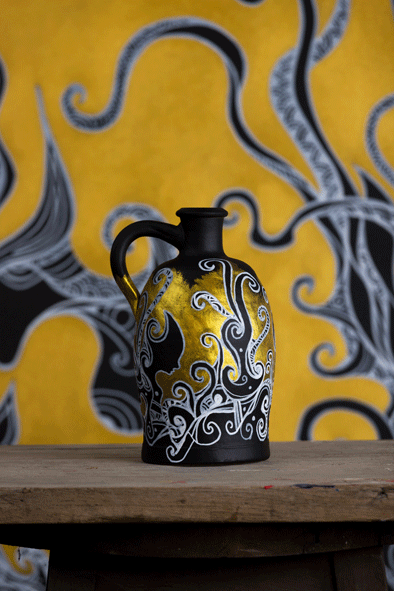 A black jug that contains a beautiful elegant hand drawn artwork with acrylic paint by artist kennetn kenisman. The gold paint gives the artwork a feeling of depth. The artwork on the object depicts a nature scene, with curls, waves, sea or nature elements and intricate details. Also silhouette figures wearing an ancient helmet. The composition is well balanced. The jug reminds of an old ancient Greek jug.